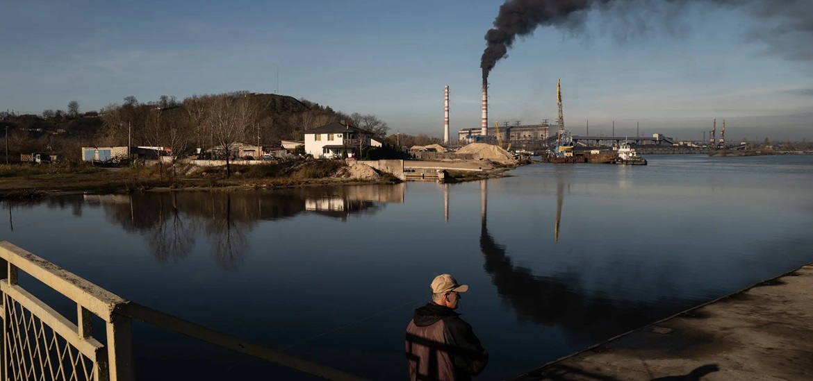 cold, cloudy winter day; older man next to a water surfice looking in the distance; black smoke rising from a factory chimney neraby