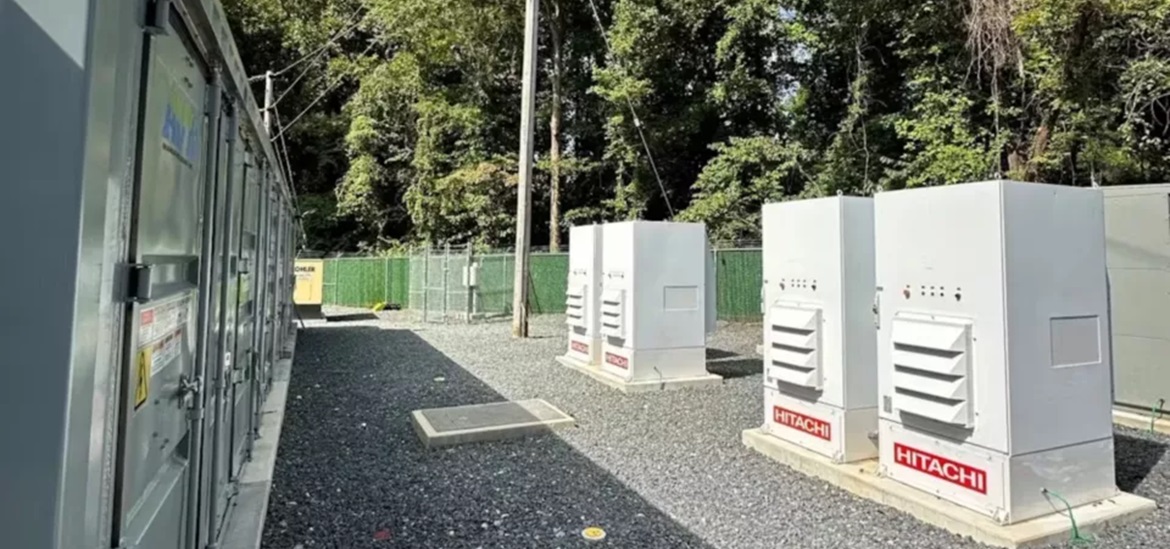 4 White BESS batteries with Hitachy logo on them as a part of a substation, surronded by fence 