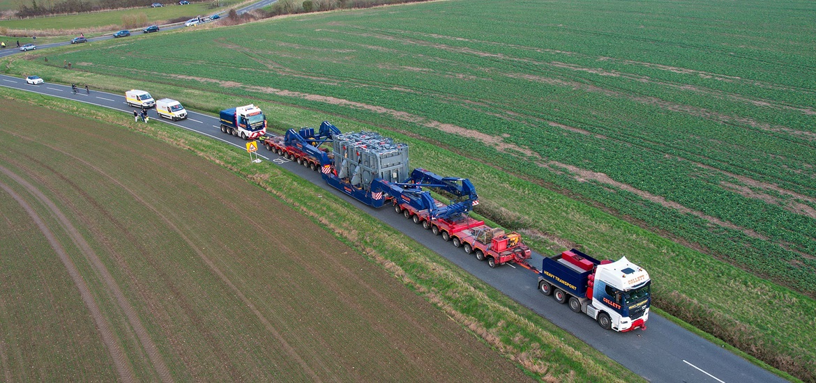 A Collett truck carrying massive transformer leading a convoy through the green fields - taken from air