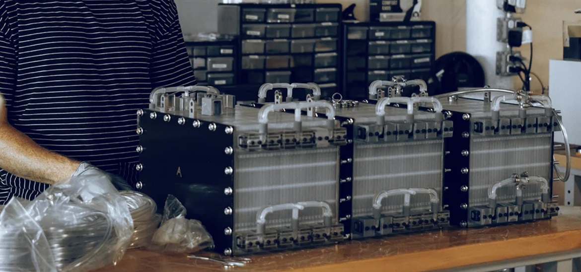 An Influit Energy technician in striped shirt stands behind awooden table bearing three rectangular prisms with a metallic shine assembling stacks of battery cells.