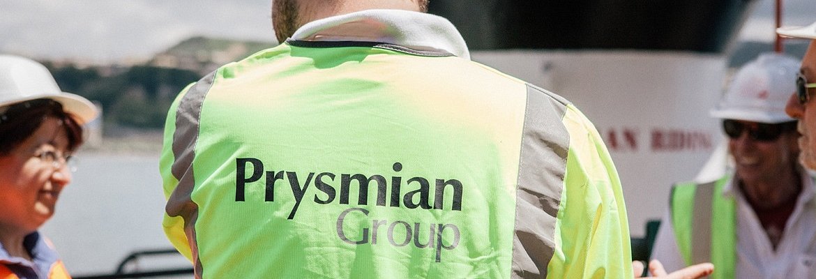 an engineer turning back to the camera with Prysmian logo on his orange vest, talking to his team
